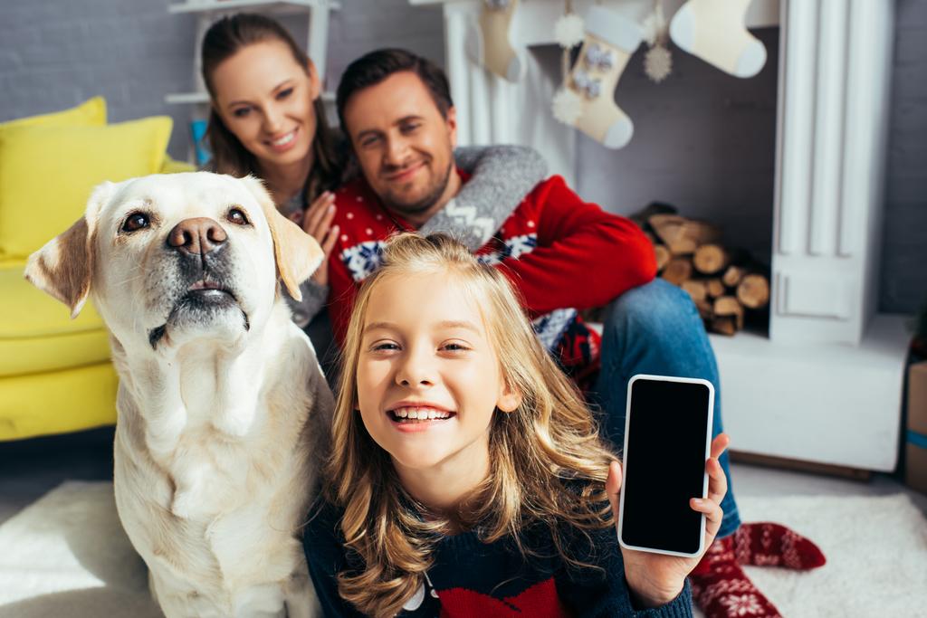 girl holding smartphone with blank screen near dog and parents on blurred background - Photo, Image