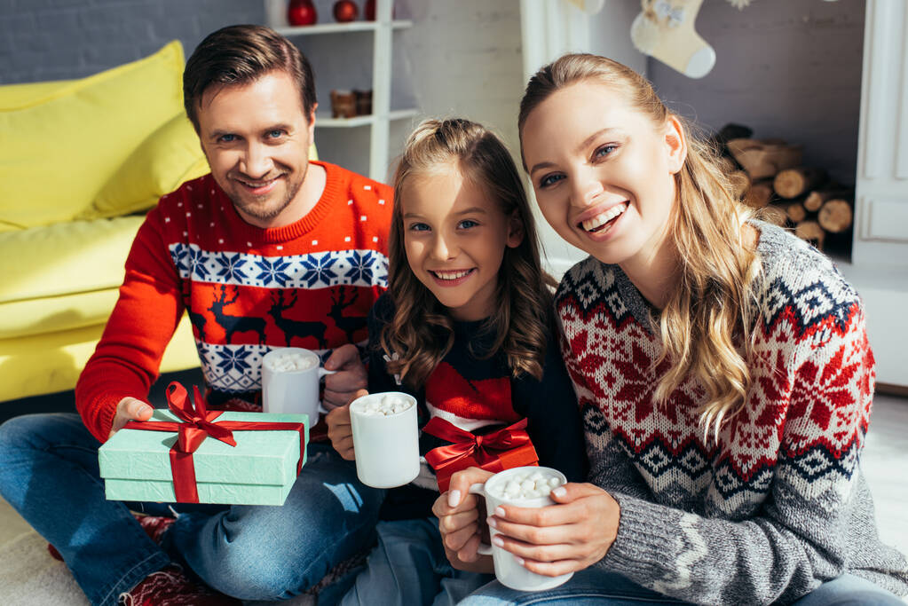 familia en suéteres sosteniendo copas con bebidas cerca presente en Navidad  - Foto, Imagen