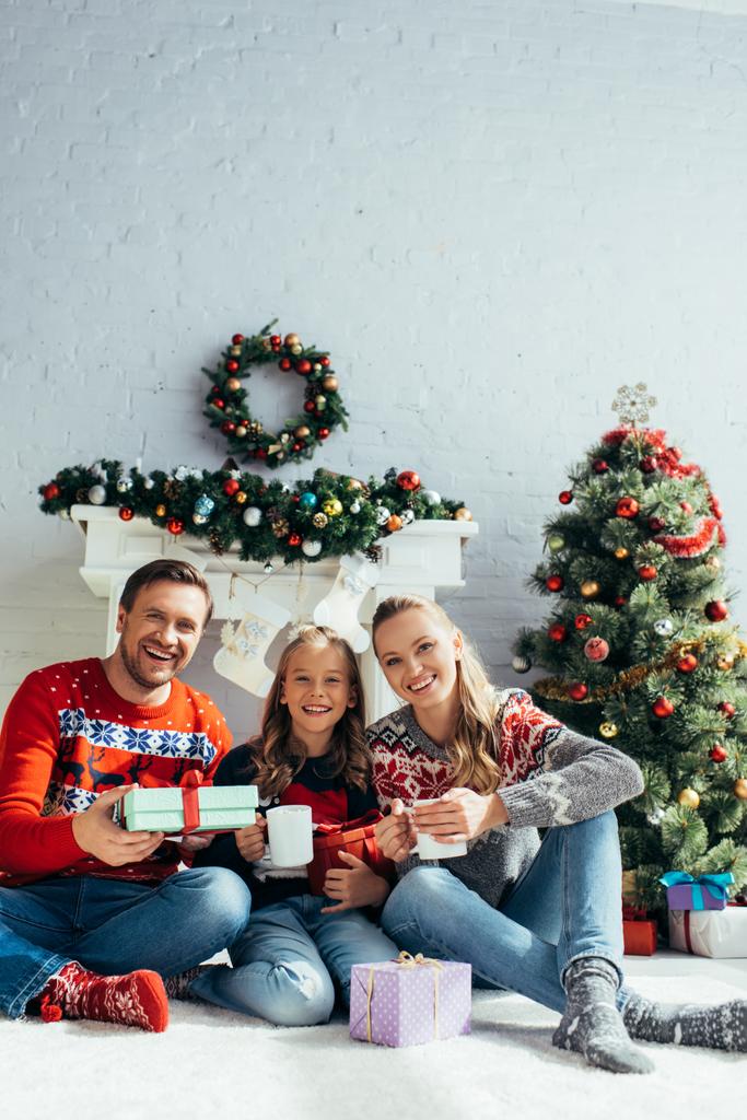 glückliche Familie hält Tassen und Geschenke in der Nähe des Weihnachtsbaums  - Foto, Bild