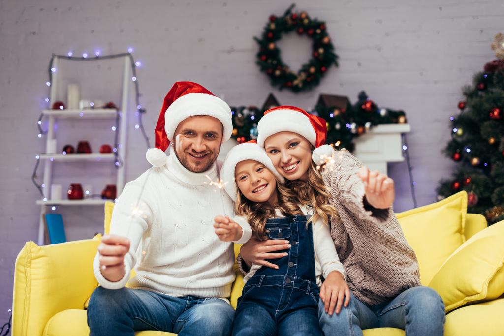 Ευτυχισμένη οικογένεια με καπέλα santa κρατώντας sparklers στο διακοσμημένο σαλόνι τα Χριστούγεννα  - Φωτογραφία, εικόνα