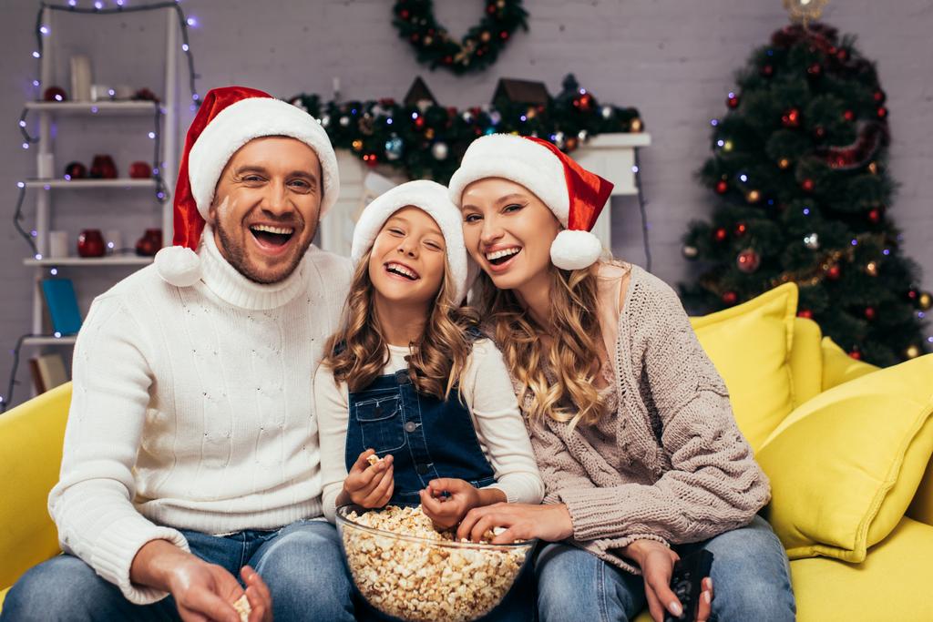 szczęśliwa rodzina w kapeluszach Mikołaja śmiejąc się i patrząc na aparat w pobliżu miski z popcornem  - Zdjęcie, obraz