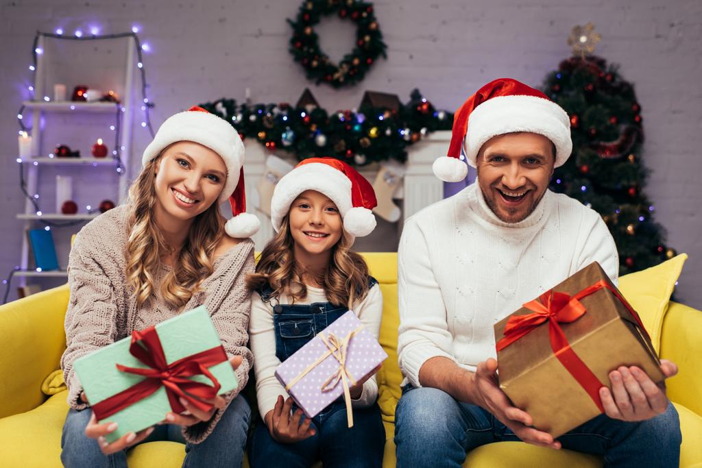 χαρούμενη οικογένεια με καπέλα santa κρατώντας δώρα στο διακοσμημένο σαλόνι τα Χριστούγεννα  - Φωτογραφία, εικόνα