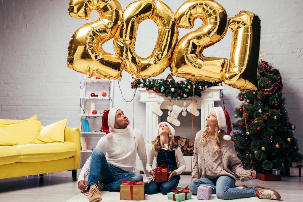 překvapená rodina v Santa klobouky sedí v blízkosti dárky a lesklé balónky s čísly 2021  - Fotografie, Obrázek