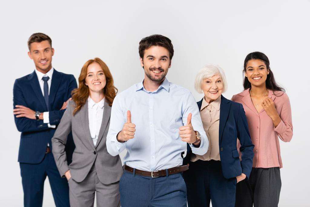 Lächelnder Geschäftsmann zeigt Daumen hoch neben fröhlichen multiethnischen Kollegen, die in grau isoliert sind - Foto, Bild