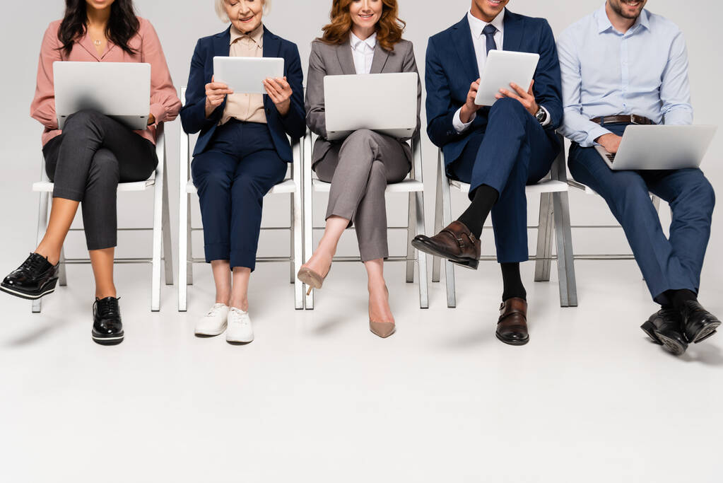 Vista recortada de empresarios multiétnicos sonrientes usando dispositivos en sillas sobre fondo gris - Foto, imagen