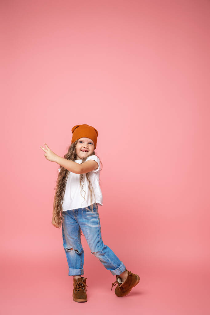 стильная маленькая девочка 6 лет с вьющимися волосами в модной шляпе и порванных джинсах стоит на розовом фоне. Показывает знак победы. ребенок улыбается беззубым ртом. Эмоциональный образ. - Фото, изображение