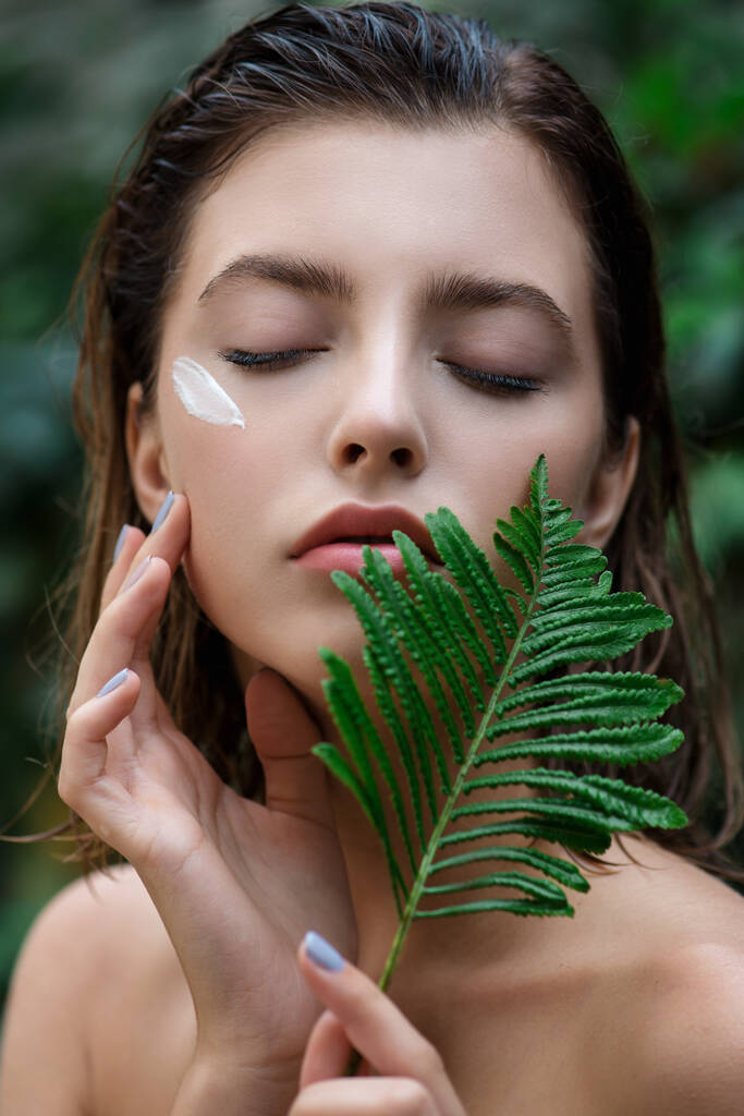 Beautiful young woman with perfect skin and natural make up posing front of plant tropical green leaves background with fern. Teen model with wet hair care of her face and body. SPA, wellness, bodycare and skincare.  Close up, selective focus. - Photo, Image