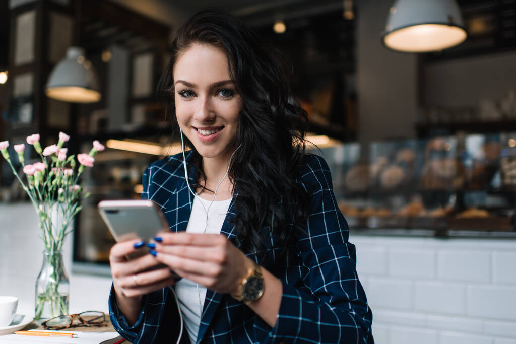 Весела жінка в елегантному вбранні та наручних годинниках сидить у затишному кафе та переглядає смартфон під час прослуховування музики на навушниках
 - Фото, зображення