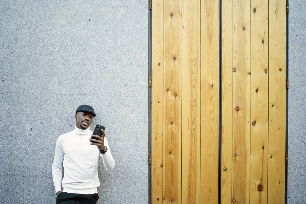 Um close-up de um homem negro usando um chapéu e uma gola alta olhando para seu telefone - Foto, Imagem