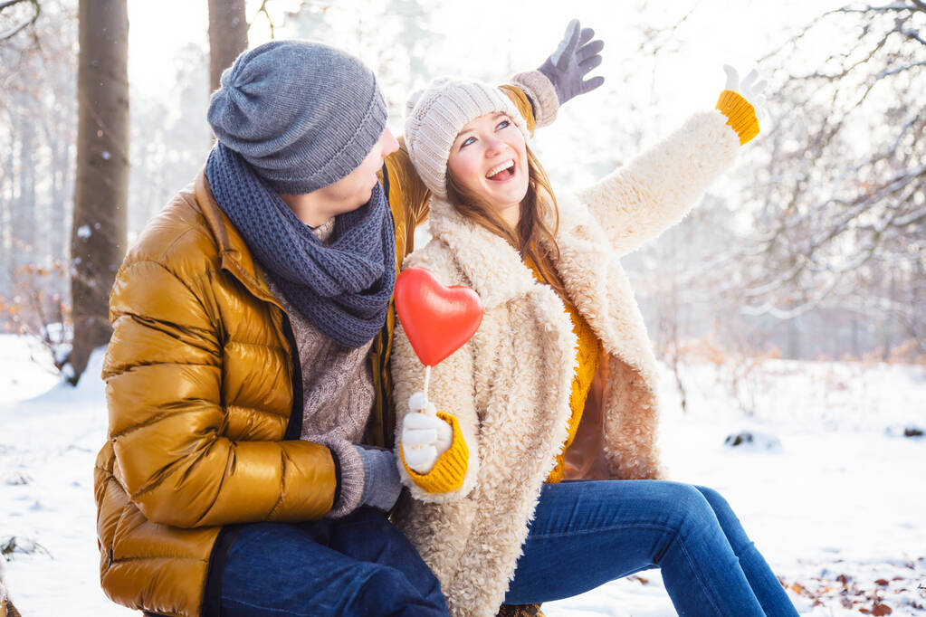 счастливая молодая пара с красным сердцем, сидя вместе в зимнем парке    - Фото, изображение