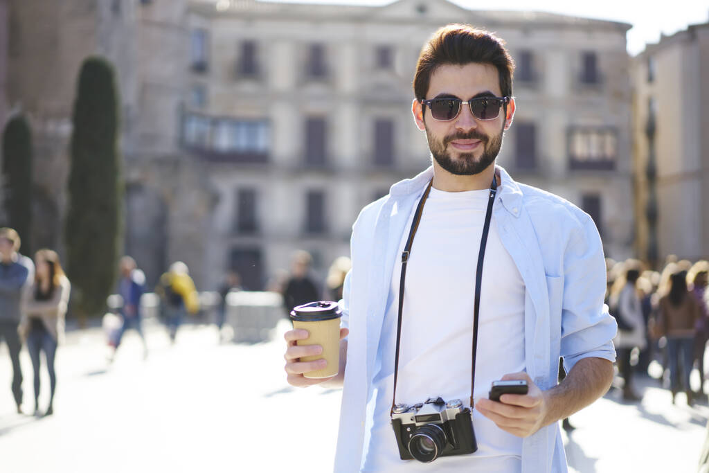 Uomo barbuto pensieroso in abito casual e occhiali da sole con fotocamera fotografica in piedi sulla strada e guardando la fotocamera mentre chatta su smartphone - Foto, immagini