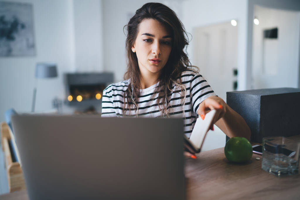 Seriöse junge Frau in lässigem Outfit sitzt am Tisch und blättert im Laptop, während sie fern arbeitet und zu Hause auf das Notizbuch schaut - Foto, Bild