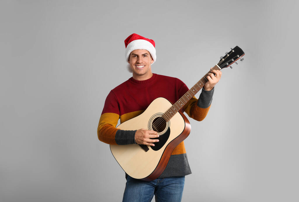 Homem de chapéu de Santa tocando guitarra acústica em fundo cinza claro. Música de Natal - Foto, Imagem
