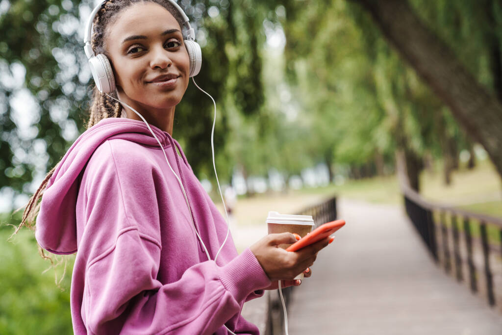 Attraente giovane donna sportiva africana che ascolta musica con le cuffie nel parco, tiene il cellulare, beve caffè da asporto - Foto, immagini