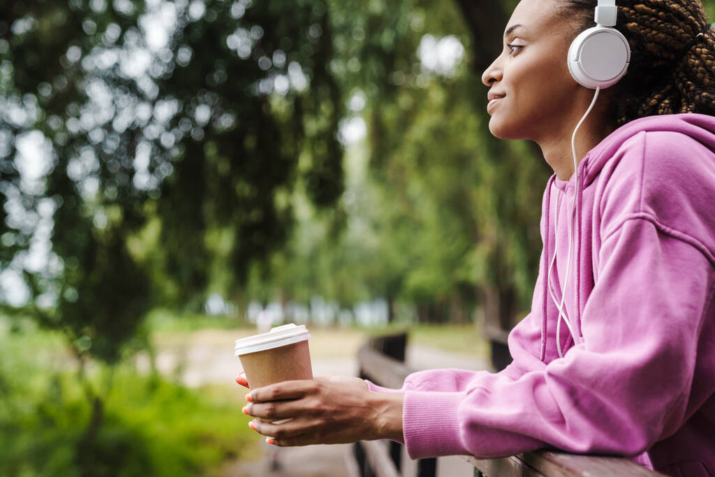 公園でヘッドフォンで音楽を聴く魅力的な若いアフリカのスポーツ女性,携帯電話を持ちます,テイクアウトコーヒーを飲みます - 写真・画像