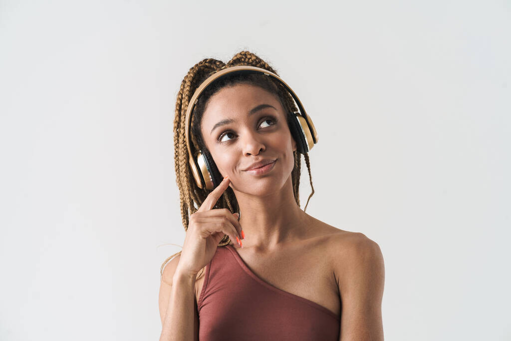 Portrait of beautiful smiling african brunette woman with dreadlocks a listening music via wireless headphones isolated on white background, looking away - 写真・画像