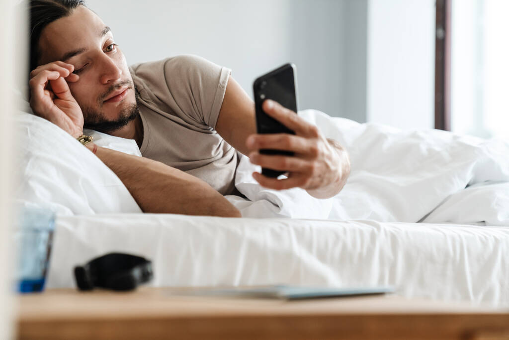 zmęczony senny młody człowiek za pomocą telefonu komórkowego podczas leżenia w łóżku rano - Zdjęcie, obraz