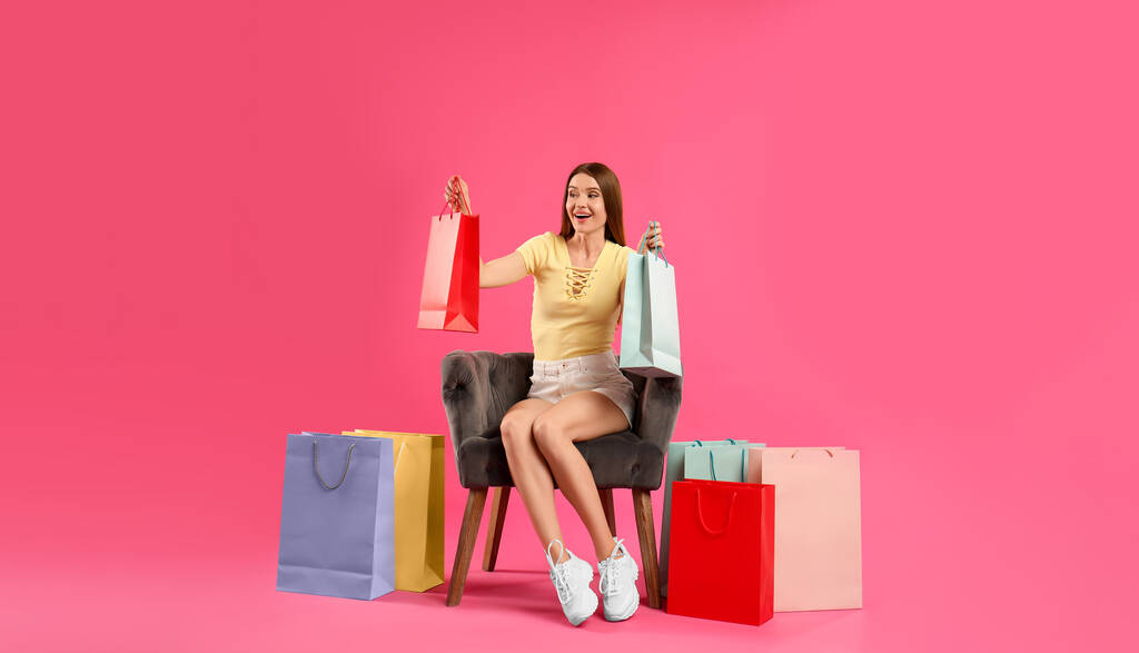 Piękna młoda kobieta z papierowymi torbami na zakupy w fotelu na różowym tle - Zdjęcie, obraz