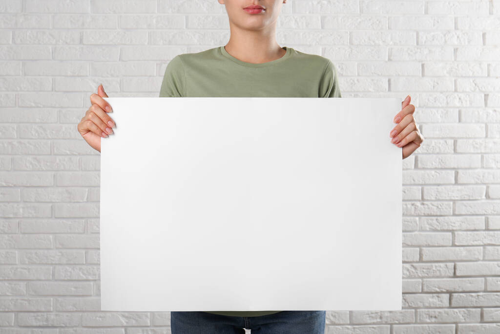 Woman holding blank poster near white brick wall, closeup. Mockup for design - Photo, Image