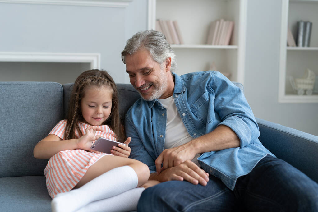 Cute girl pomóc ojcu pokazać coś na smartfonie, córeczka i tata siedzieć na kanapie trzymać telefon komórkowy - Zdjęcie, obraz