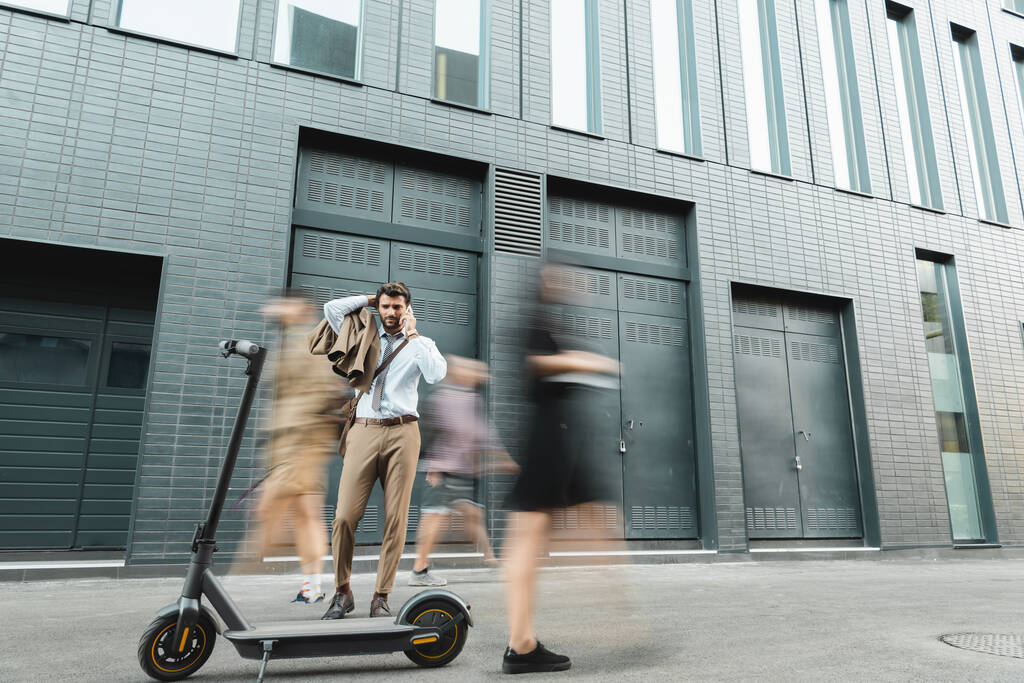 Takım elbiseli bir işadamı e-scooter ve insanların yanında dururken akıllı telefondan konuşuyor, hareket bulanıklığı - Fotoğraf, Görsel
