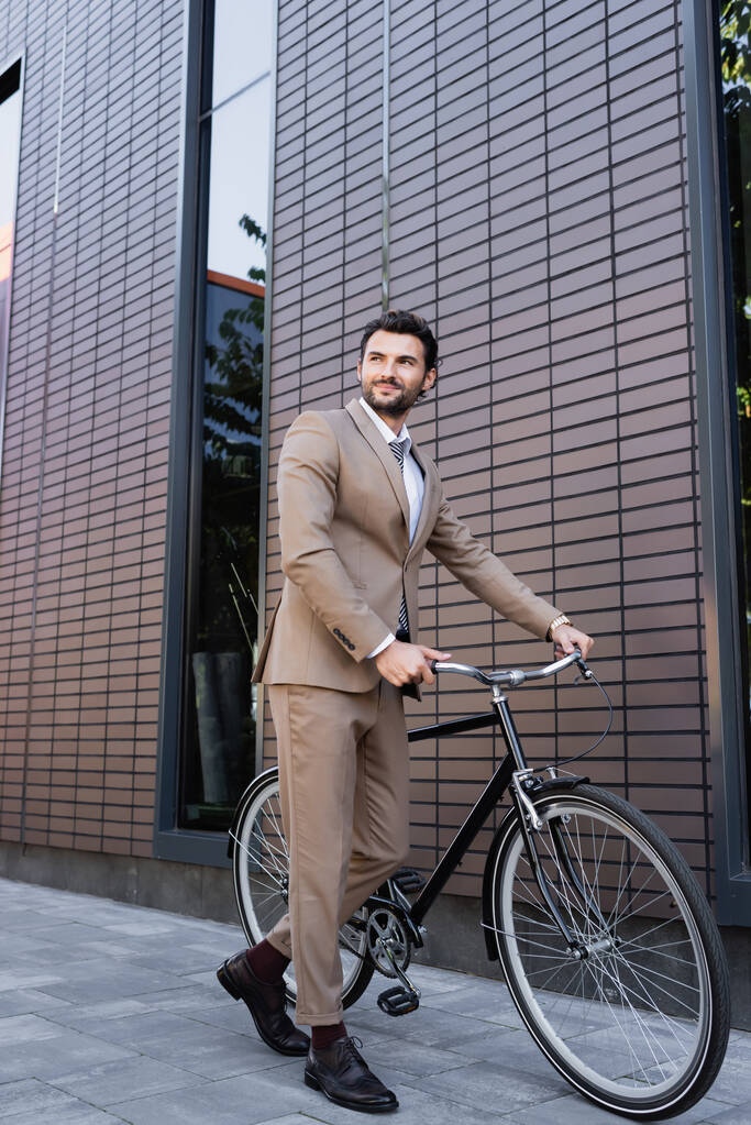 pełna długość szczęśliwy biznesmen w garniturze spacery z rowerem w pobliżu budynku  - Zdjęcie, obraz