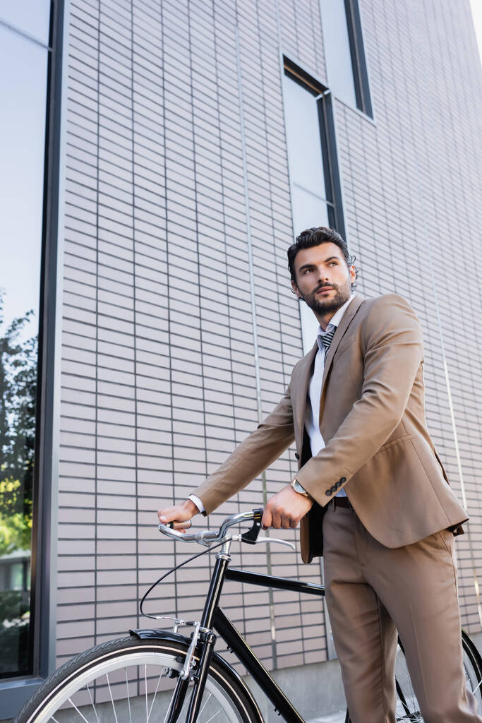 низкий угол зрения бородатый бизнесмен в костюме, стоящий с велосипедом, глядя в сторону рядом со зданием  - Фото, изображение