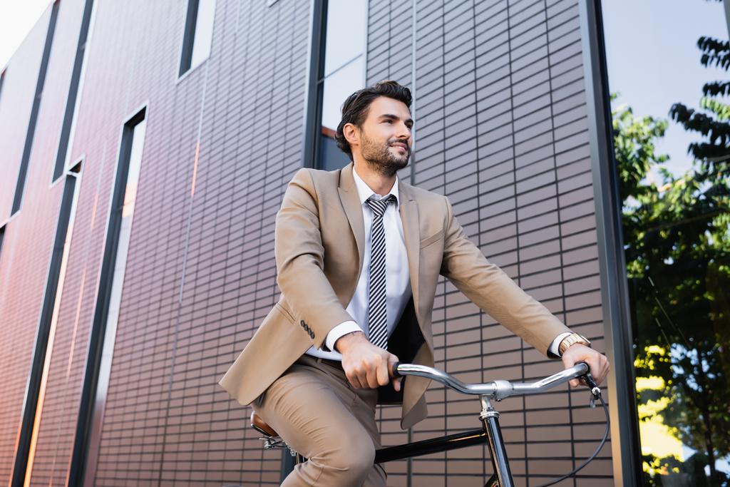 Tiefansicht eines bärtigen Mannes im Anzug, der Fahrrad fährt, lächelt und in der Nähe des Gebäudes wegschaut  - Foto, Bild