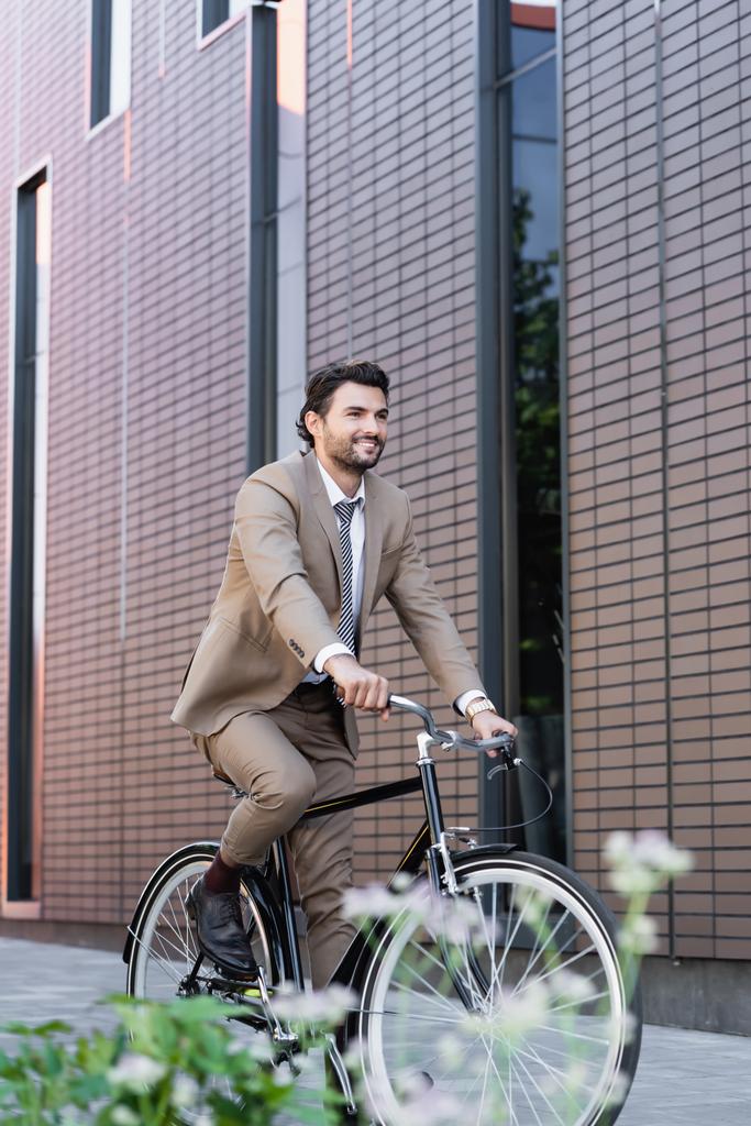 hombre feliz en traje de montar en bicicleta y sonriendo cerca de la construcción y la planta en primer plano borrosa  - Foto, Imagen