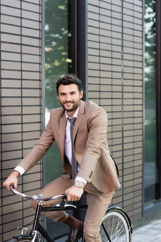 homme d'affaires heureux en costume à vélo et regardant la caméra près du bâtiment  - Photo, image