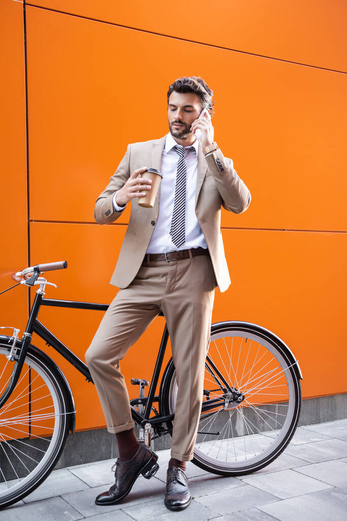 comprimento total de homem de negócios falando no smartphone e segurando copo de papel perto de bicicleta e construção com paredes laranja - Foto, Imagem