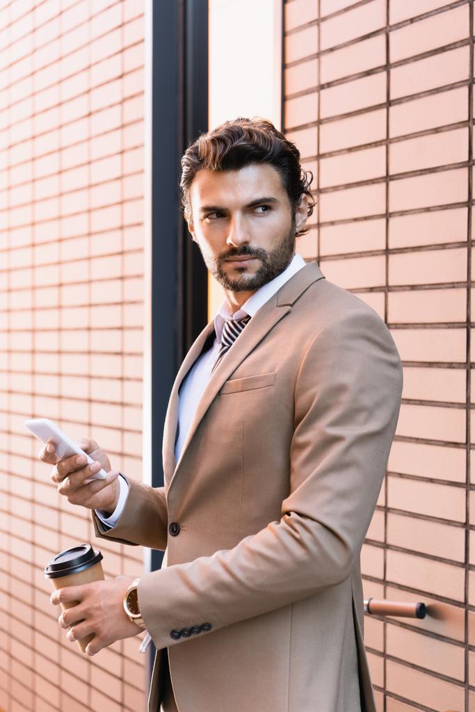hombre de negocios barbudo sosteniendo teléfono inteligente y taza de papel mientras está de pie cerca del edificio  - Foto, Imagen