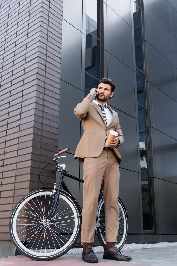 niski kąt widzenia biznesmena mówiącego na smartfonie i trzymającego papierową filiżankę w pobliżu roweru i budynku  - Zdjęcie, obraz