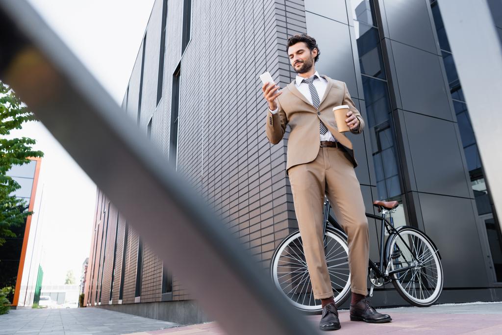Tiefansicht des gut gelaunten Geschäftsmannes, der aufs Smartphone schaut und Pappbecher neben Fahrrad hält, und verschwommener Vordergrund  - Foto, Bild