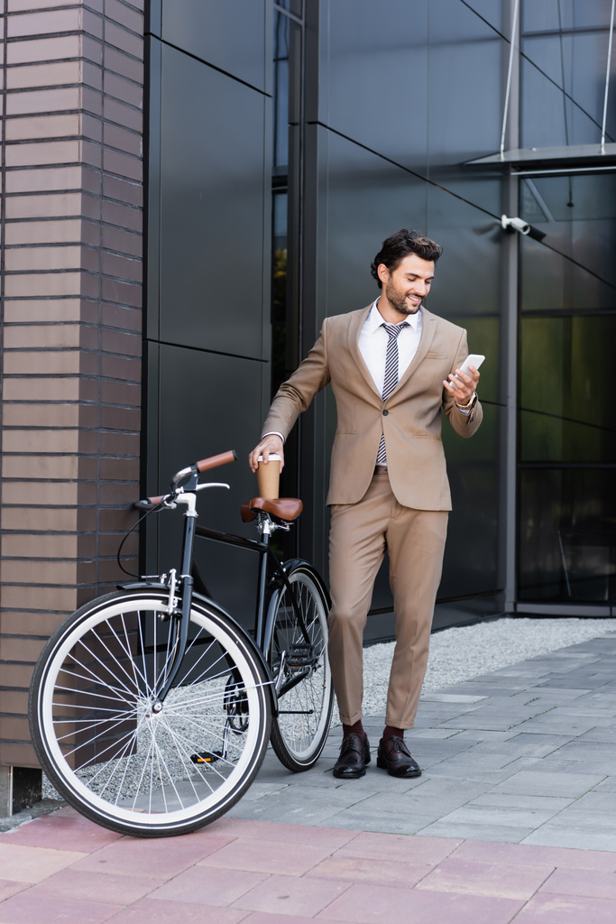повна довжина веселого бізнесмена, який тримає смартфон і каву, щоб піти поблизу велосипеда, стоячи біля будівлі
  - Фото, зображення