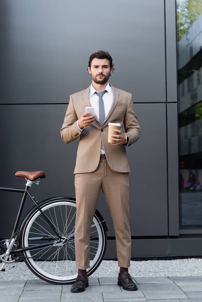 Sakallı bir işadamı elinde kahve, bisiklet yanında akıllı telefon tutuyor.  - Fotoğraf, Görsel