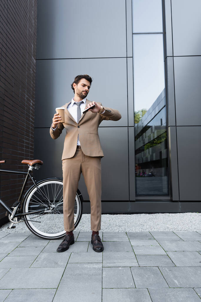 plná délka vousatý podnikatel v obleku drží kávu jít a smartphone při pohledu na hodinky v blízkosti kola  - Fotografie, Obrázek