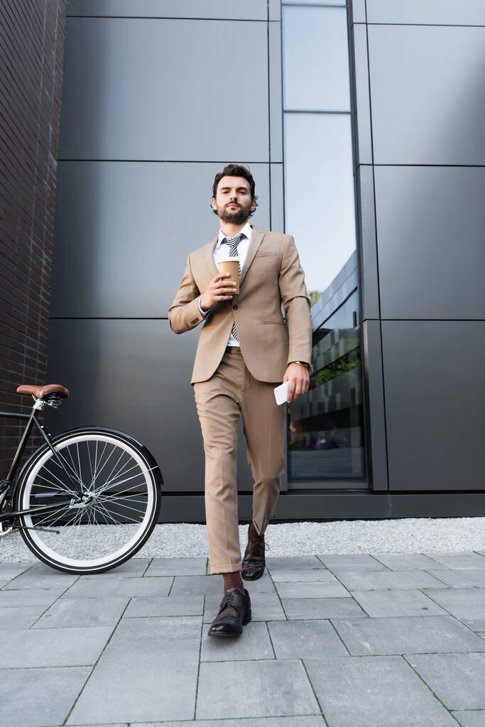 longitud completa de hombre de negocios barbudo en traje sosteniendo café para ir y teléfono inteligente mientras camina cerca de la bicicleta  - Foto, imagen