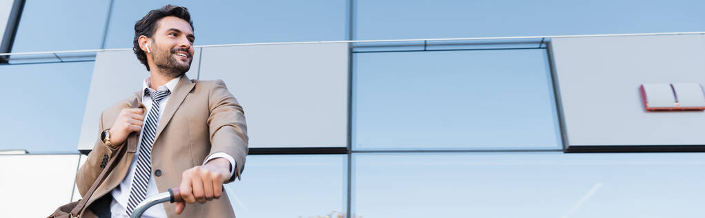 visão de baixo ângulo de empresário alegre em fones de ouvido sem fio enquanto de pé com saco perto de bicicleta e construção, banner - Foto, Imagem
