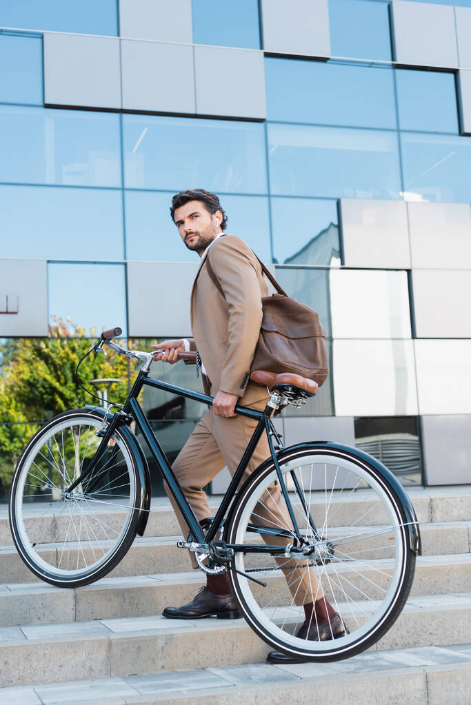 hombre de negocios serio en auriculares inalámbricos caminando en escaleras con bicicleta  - Foto, Imagen