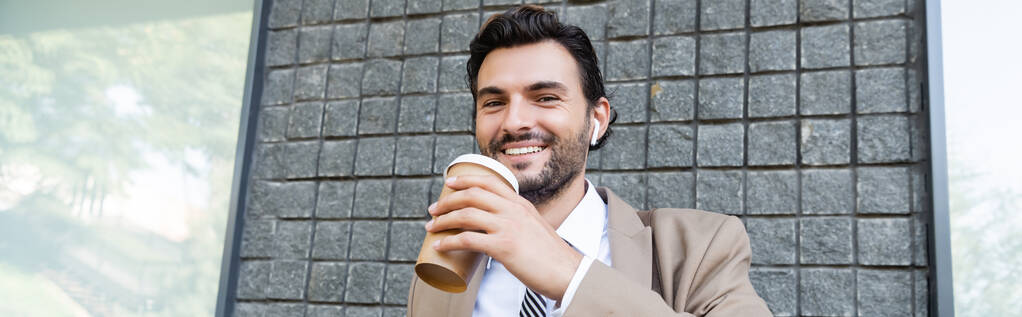 счастливый бизнесмен в беспроводных наушниках держа бумажную чашку возле здания, баннер - Фото, изображение