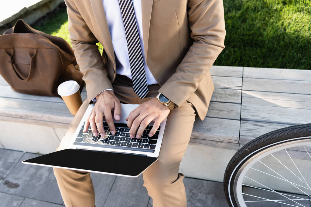 vista parcial del hombre de negocios utilizando el ordenador portátil cerca de la taza de papel y sentado en el banco  - Foto, Imagen