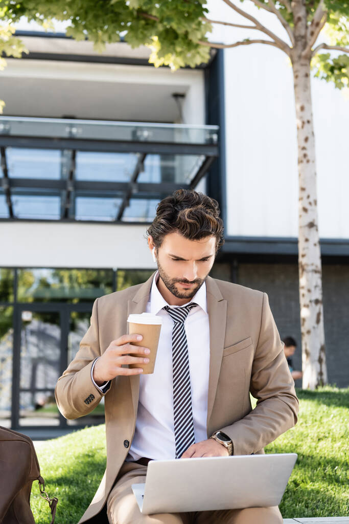liikemies langattomissa kuulokkeissa ja puku tilalla kahvia mennä ja käyttää kannettavaa tietokonetta istuessaan penkillä  - Valokuva, kuva