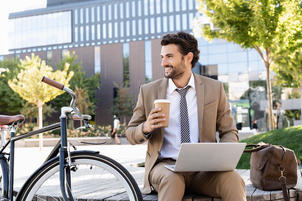 homme d'affaires heureux en costume à l'aide d'un ordinateur portable et tenant tasse en papier tout en étant assis sur le banc près de vélo - Photo, image