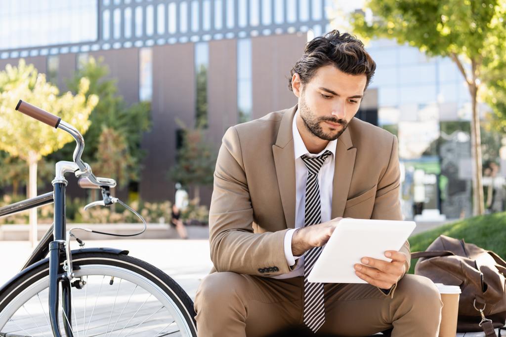 homme d'affaires en costume tenant tablette numérique tout en étant assis sur le banc près de vélo - Photo, image