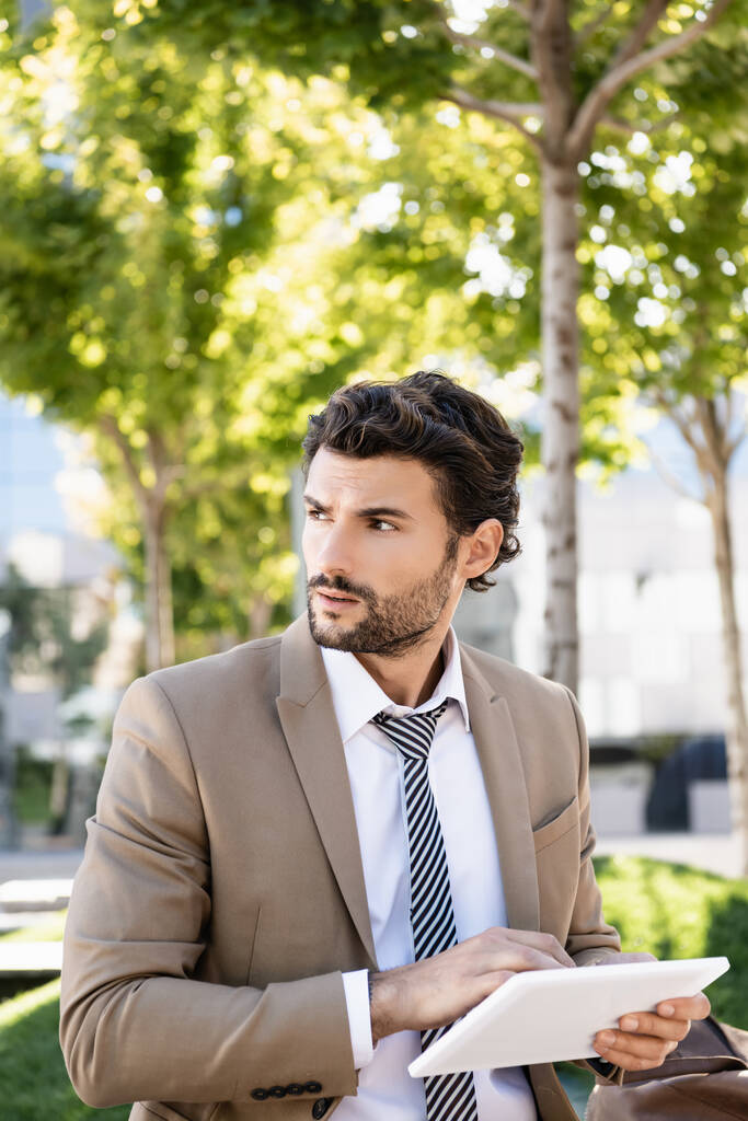 бородатый бизнесмен в костюме держит цифровой планшет, сидя на скамейке  - Фото, изображение