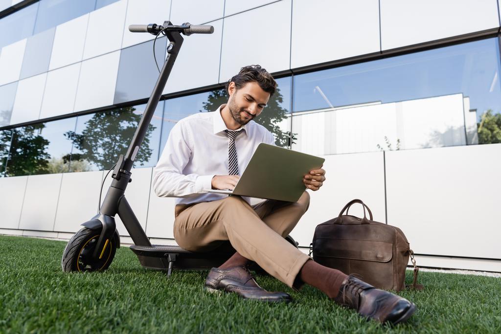 hombre de negocios sonriente en ropa formal sentado con el ordenador portátil cerca de e-scooter en la hierba - Foto, imagen