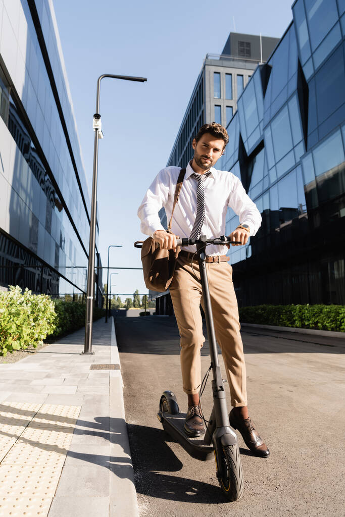 В полный рост бизнесмен в формальной одежде с кожаной сумкой ездит на электроскутере  - Фото, изображение