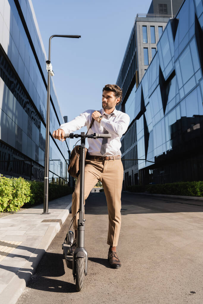 longitud completa de hombre de negocios en desgaste formal ajustando bolsa de cuero mientras monta scooter eléctrico  - Foto, imagen