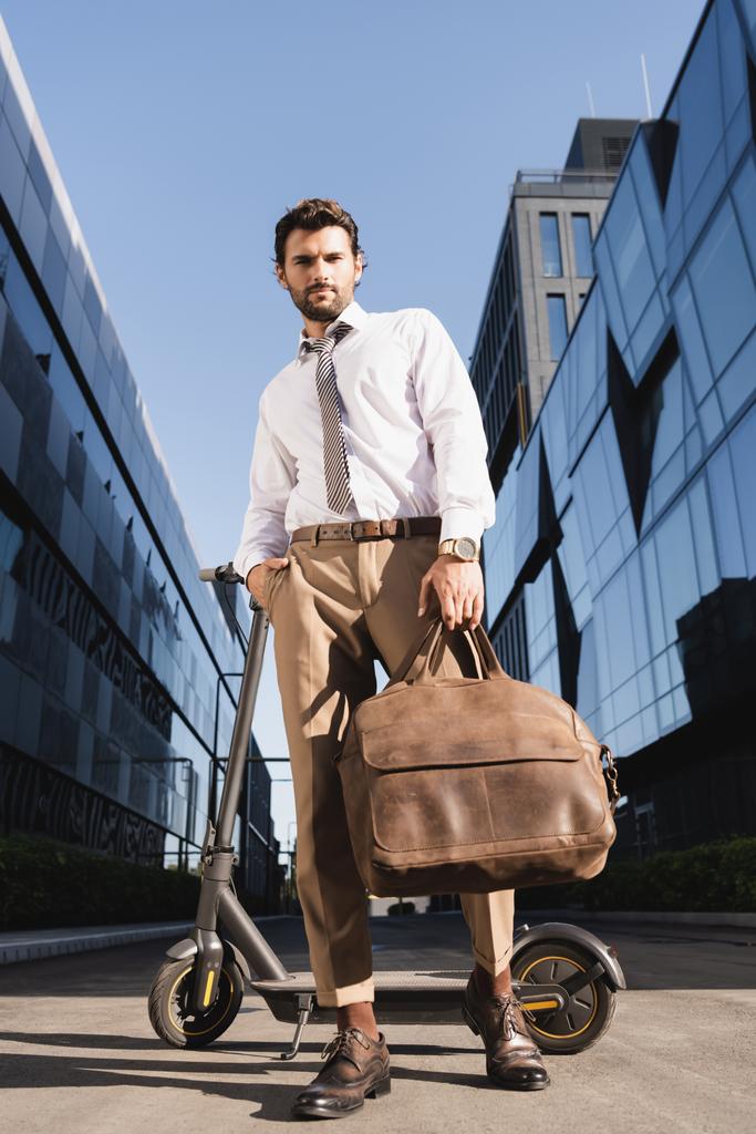 low angle view of bearded businessman in formal wear holding leather bag while standing with hand in pocket near electric scooter  - Photo, Image